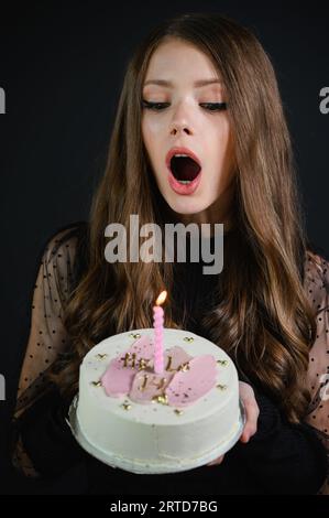 Schönes, glückliches Mädchen, das ein schwarzes Kleid trägt und eine Kerze auf einem Geburtstagskuchen ausbläst. Kuchen mit der Aufschrift Hallo 14. Stockfoto