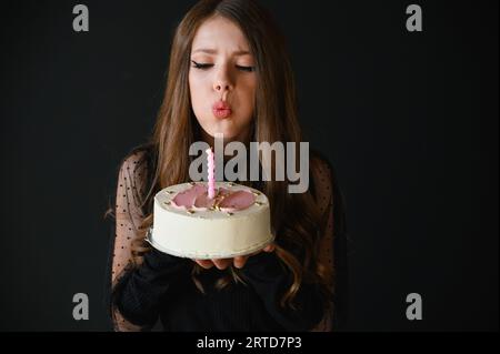 Schönes, glückliches Mädchen im schwarzen Kleid, das eine Kerze auf dem Geburtstagskuchen ausbläst. Schwarzer Hintergrund. Stockfoto