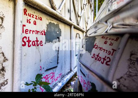 Das Graffiti "Ruzzia ist ein Terrorstaat" ist in Rot auf einer weiß gestrichenen Metallwand in der Revaz-Tabukashvili-Straße in Tiflis, Georgien, geschrieben. Die Karosserie eines geparkten Autos spiegelt die Schrift wider. Die Schreibweise "Ruzzia" spielt auf das Z an, das Kennzeichen der russischen Truppen bei der Invasion der Ukraine im Jahr 2022. Daneben bedeckt ein grauer Farbfleck ältere Schriftzüge, und am Fuß der Wand wächst eine Pflanze. Im Jahr 2023 prägten Graffiti gegen Russland und für engere Beziehungen zu Europa das Straßenbild von Tiflis. Im August 2008 führte Russland Krieg gegen das Land. Stockfoto
