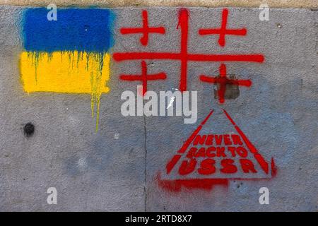 Die schablonierten Worte „Niemals zurück in die USSR“ erscheinen in einem roten Dreieck auf einer grauen Hauswand in Tiflis, Georgien. Darüber befinden sich die blauen und gelben Farben der ukrainischen Flagge und das rote Kreuzmotiv der georgischen Nationalflagge. Die gelbe Farbe ist in Tropfen heruntergelaufen. Im Jahr 2023 prägten Graffiti gegen Russland und für engere Beziehungen zu Europa die Straßenlandschaft von Tiflis. Stockfoto
