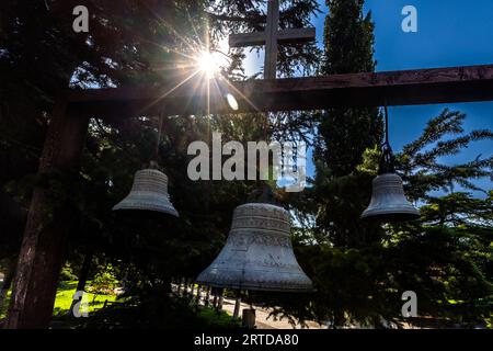 In Georgien hängen die Kirchenglocken normalerweise außerhalb der Kirchen. Vor der Metekhi-Kirche der Geburt der Mutter Gottes in Tiflis, Georgien, hängen sie an einem Holzbau. Oben auf der Struktur befindet sich ein Kreuz. Sonnenlicht scheint durch die Bäume. Das Kirchengebäude stand ursprünglich in der Mitte der königlichen Residenz Stockfoto