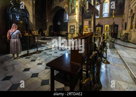 Eine Frau mit Kopftuch steht in der Kathedrale von Sioni in Tiflis, Georgia. Sie sieht sich einen Bereich mit Kerzen und Ikonen an. Das Innere ist mit Wandmalereien und Marmorboden dekoriert. Tiflis, Georgien Stockfoto