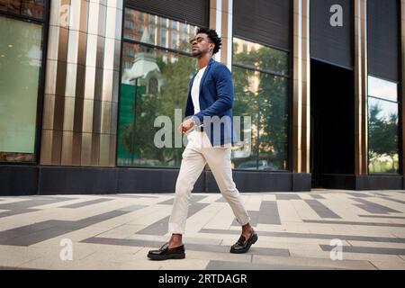 Ein Mann mit seitlichem Blick geht draußen spazieren und trägt trendige Geschäftskleidung, blaue Jacke, weiße Hosen und Slipper. Stilvolles, modisches männliches Modell, Stre Stockfoto