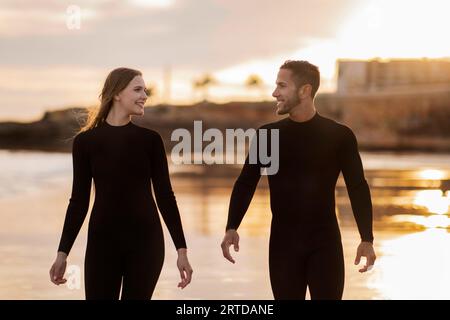 Glückliches Junges Paar, Das Wetsuits Trägt, Die Am Strand Zur Sonnenuntergangszeit Spazieren Stockfoto