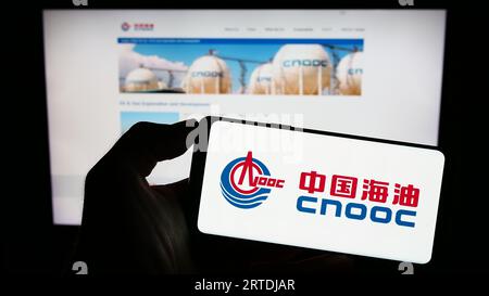 Person, die ein Mobiltelefon mit dem Logo der China National Offshore Oil Corporation (CNOOC) auf dem Bildschirm vor der Webseite hält. Konzentrieren Sie sich auf das Display des Telefons. Stockfoto