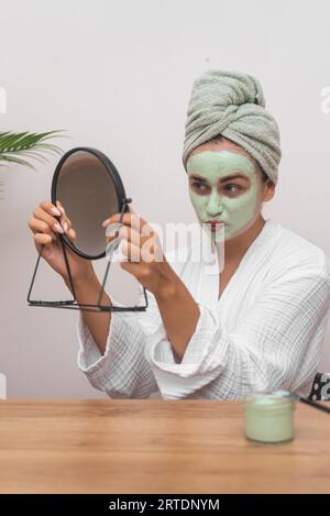 Frau verwöhnt den Tag, trägt grüne Tonmaske auf und lächelt über ihr strahlendes Spiegelbild im Spiegel. Stockfoto