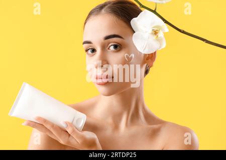 Junge Frau mit Tube Naturkosmetik und Orchideenblüten auf gelbem Hintergrund Stockfoto