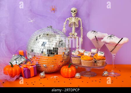 Halloween-Party-Komposition mit Cupcakes, Disco-Ball, Getränken und Dekorationen auf orangenem Tisch vor lila Hintergrund Stockfoto