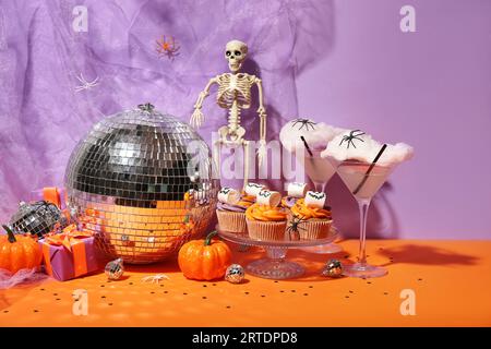 Halloween-Party-Komposition mit Cupcakes, Disco-Ball, Getränken und Dekorationen auf orangenem Tisch vor lila Hintergrund Stockfoto