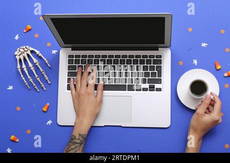 Weibliche Hände mit Laptop, Tasse Kaffee und Halloween-Deko auf blauem Hintergrund Stockfoto