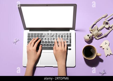 Frauenhände mit Laptop, Tasse Kaffee und Halloween-Deko auf fliedernem Hintergrund Stockfoto