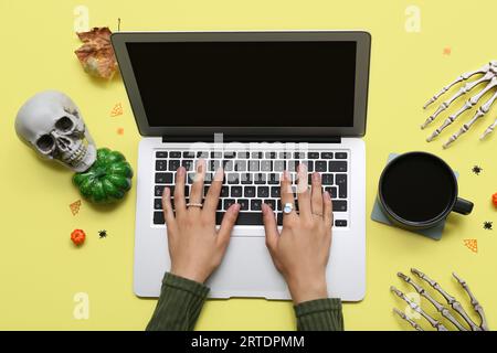 Weibliche Hände mit Laptop, Tasse Kaffee und Halloween-Deko auf gelbem Hintergrund Stockfoto