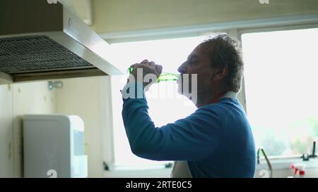 Älterer Mann trinkt Bier aus der Flasche, während er Essen mit dem Küchenherd kocht, Schürze trägt ältere Person trinkt alkoholisches Getränk während der Zubereitung der Mahlzeit Stockfoto