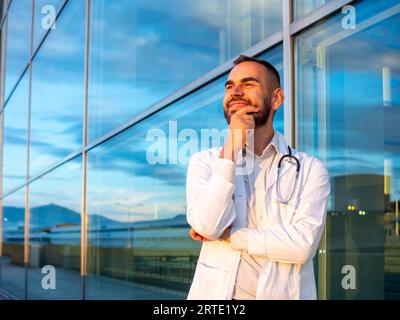 Junger männlicher Arzt mit einem Stethoskop am Hals lächelnd vor einem Krankenhaus Stockfoto