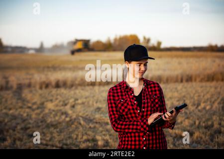 Porträt einer jungen Landwirtschaftsfrau, die zur Erntezeit auf einem Getreidefeld steht und fortschrittliche landwirtschaftliche Softwaretechnologien auf einem Pad verwendet, während ein Kombi... Stockfoto