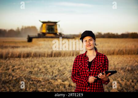 Porträt einer jungen Landwirtschaftsfrau, die zur Erntezeit auf einem Getreidefeld steht und fortschrittliche landwirtschaftliche Softwaretechnologien auf einem Pad verwendet, während ein Kombi... Stockfoto