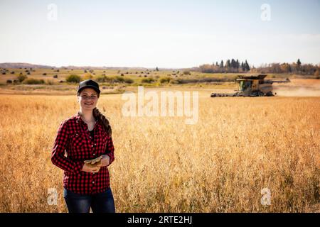 Porträt einer jungen Landwirtschaftsfrau, die zur Erntezeit auf einem Mischkornfeld steht, während ein Mähdrescher im Hintergrund arbeitet Stockfoto