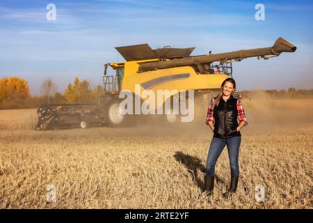 Porträt einer Reifen Landwirtschaftsfrau, die auf einem Getreidefeld steht und während der Ernte für die Kamera posiert, während ein Mähdrescher bei Sonnenuntergang im Hintergrund arbeitet Stockfoto