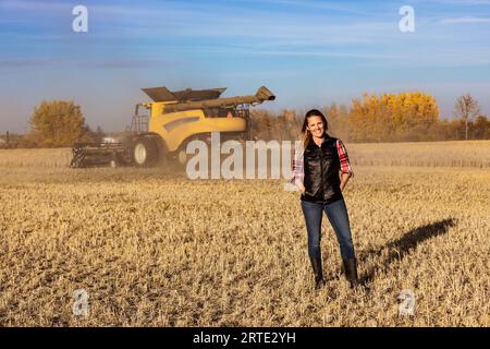 Porträt einer Reifen Landwirtschaftsfrau, die auf einem Getreidefeld steht und während der Ernte für die Kamera posiert, während ein Mähdrescher bei Sonnenuntergang im Hintergrund arbeitet Stockfoto