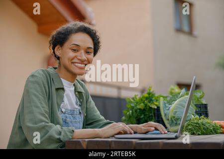 Lächelnde Frau Gemüsehausbesitzer, der am Laptop arbeitet, mit Bestellungen im Hintergrund des Gebäudes Stockfoto