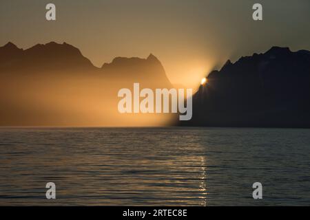 Goldenes Licht des Sonnenaufgangs, das um die Silhouetten der Berge schaut, die sich im Wasser der westlichen Fjorde von Kalaallit Nunaat spiegeln Stockfoto