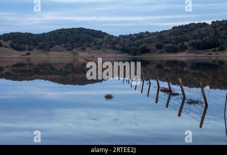 Ein Holz- und Drahtzaun zieht sich quer durch und ein großes Gewässer. Das Wasser ist blau. Es gibt Hügel hinter dem Wasser. Der Himmel ist hellblau und bewölkt Stockfoto