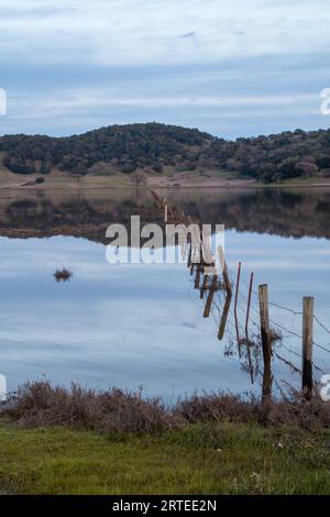 Ein Holz- und Drahtzaun zieht sich quer durch und ein großes Gewässer. Das Wasser ist blau. Es gibt Hügel hinter dem Wasser. Der Himmel ist hellblau und bewölkt Stockfoto