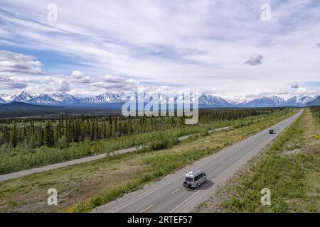 Aus der Vogelperspektive auf einigen Wohnmobilen, die durch die bewaldete Landschaft entlang des Alaska Highway fahren, mit fantastischem Blick auf die schneebedeckten Bergketten auf... Stockfoto