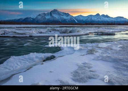 Die Sonne geht hinter Pioneer und Twin Peaks und der Chugach Mountain Range in einer Winternacht unter, mit einem seidenglatten Matanuska River, gesäumt von Eis... Stockfoto