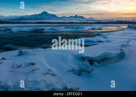 Die Sonne geht hinter Pioneer und Twin Peaks und der Chugach Mountain Range in einer Winternacht unter, mit einem seidenglatten Matanuska River, gesäumt von Eis... Stockfoto