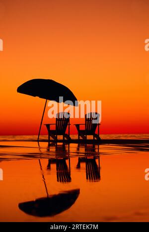 Dramatische Sonnenuntergangsszene mit zwei Stühlen und einem Sonnenschirm am Strand; Seaside, Florida, Vereinigte Staaten von Amerika Stockfoto