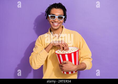 Foto eines jungen Mannes trägt gelbe Hemd halten Popcorn Eimer Essen leckere Chips 3D Film Guardians Galaxie isoliert auf violettem Hintergrund Stockfoto