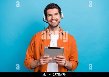 Porträt eines gutaussehenden positiven Guys, eines Zahnlächelhalters, der ein Tablet verwendet, ein kabelgebundenes Headset, das Kunden isoliert auf blauem Hintergrund kommuniziert Stockfoto