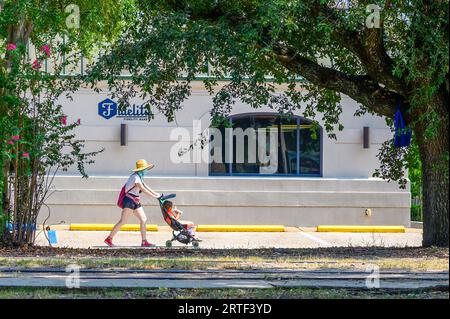 NEW ORLEANS, LA, USA: 28. AUGUST 2023: Frau mit Gesichtsmaske schiebt einen Kinderwagen mit einem Kind an einem Bankgebäude in der South Carrollton Avenue vorbei Stockfoto