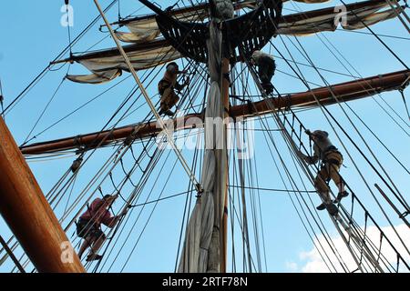 Segler werden in Silhouette vor dem blauen Himmel gesehen, während sie die Takelage eines großen Schiffes erklimmen, um die Segel zu entfalten, um den aufsteigenden Wind zu fangen Stockfoto