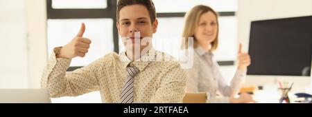 Zwei Manager sitzen im Büro an ihrem Schreibtisch und zeigen Daumen-hoch-Geste. Stockfoto