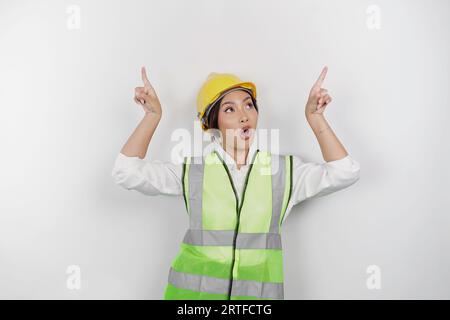 Eine schockierte asiatische Frau, die einen Schutzhelm und eine Weste trägt und auf einen Kopierraum über ihr zeigt, der durch einen weißen Hintergrund isoliert ist. Das Arbeitszeitkonzept. Stockfoto