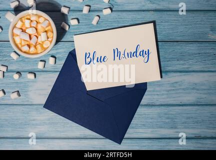 BLAUER MONTAGSTEXT mit blauem Umschlag mit weißer Tasse Kaffee und Marshmallows auf hölzernem Hintergrund. Der deprimierendste Tag des Jahres im Januar. Tag Selbstmord und Depression Motivationszeichen. Draufsicht, flach Stockfoto