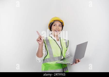 Eine lächelnde asiatische Frau arbeitet mit einem Schutzhelm und einer Weste, zeigt auf einen Kopierraum über ihr und hält ihren Laptop, isoliert durch einen weißen Hintergrund. Labor Stockfoto