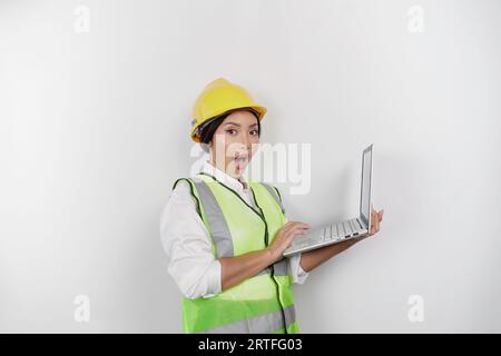 Ein Porträt einer schockierten asiatischen Frau, die einen Schutzhelm und eine Weste trägt, während sie ihren Laptop mit weit geöffnetem Mund hält, isoliert von einem weißen Backgrou Stockfoto