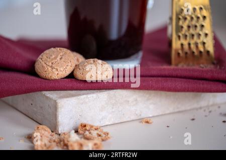 Amaretto-Kekse neben einer Kaffeetasse aus Glas auf einer roten Serviette und einem Marmortablett. Eine kleine Goldreibe im Hintergrund. Hochwertige Fotos. Stockfoto
