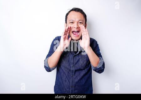 Junger, gutaussehender Mann, der ein blaues Hemd trägt, schreit und schreit laut mit einer Hand auf dem Mund. Kommunikationskonzept. Stockfoto