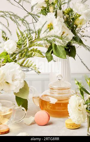 Ästhetische Teezeit, Teekanne, Kräutertee und Macarondesserts im biophilen Innenraum unter weißen Blüten. Femininer, ruhiger Lebensstil. Stockfoto