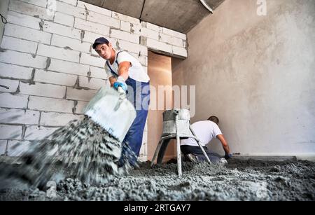 Männlicher Arbeiter mit Schaufel beim Schaufeln von Sand-Zement-Mischungen im Bau. Mann, der Bodenbodenmaterial vorbereitet, während er in der Nähe der Betonbodenmischmaschine steht. Stockfoto