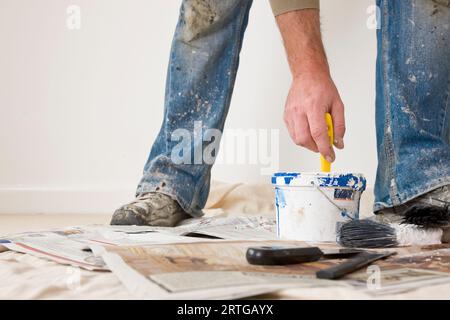 Nahaufnahme der Beine eines Mannes, der Schuhe und Jeans trägt, die mit Farbe bedeckt sind, während seine Hand einen Pinsel taucht Stockfoto