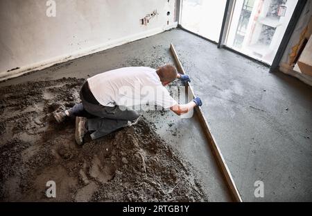 Männlicher Bauarbeiter, der Estrichschiene auf den mit Sand-Zement-Gemisch bedeckten Boden legt. Mann glättet und ebnet die Oberfläche mit dem Lineal beim Verputzen des Bodens in der Wohnung. Stockfoto