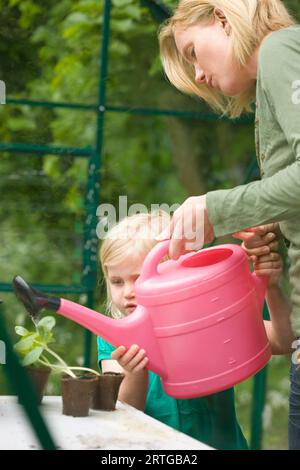 Junge blonde Mädchen hilft Frau Sämling Töpfe im Gewächshaus mit rosa Gießkanne gießen Stockfoto