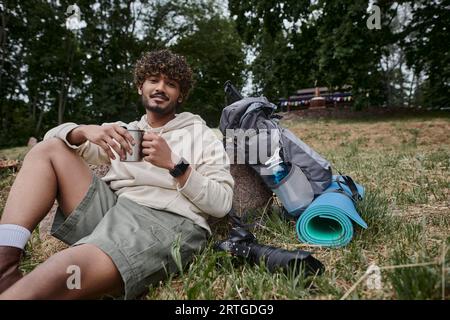 indischer Mann, der Thermosbecher in der Hand hält und neben Kamera und Rucksack mit Reiseausrüstung sitzt, Tourist Stockfoto