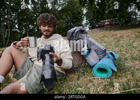 Glücklicher indischer Mann, der Thermosbecher hält und Fotos mit der Kamera betrachtet, Tourist in der Nähe von Reiseausrüstung Stockfoto