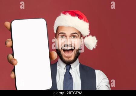 Glücklicher, erfolgreicher Geschäftsmann in rotem Weihnachtsmannhut zeigt Smartphone mit leerem weißen Bildschirm für die neue App Stockfoto